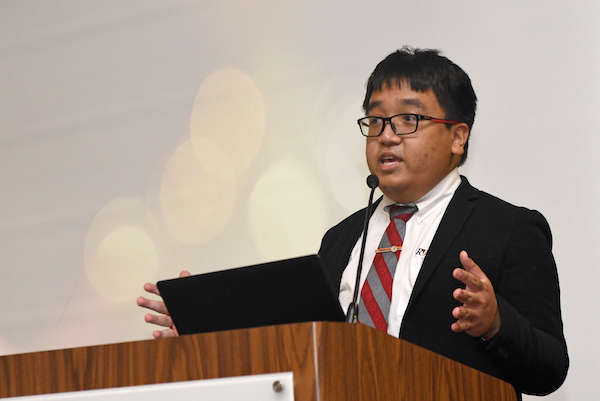 Soup's in a black suit and a maroon-grey tie standing behind a microphone and in front of a white screen